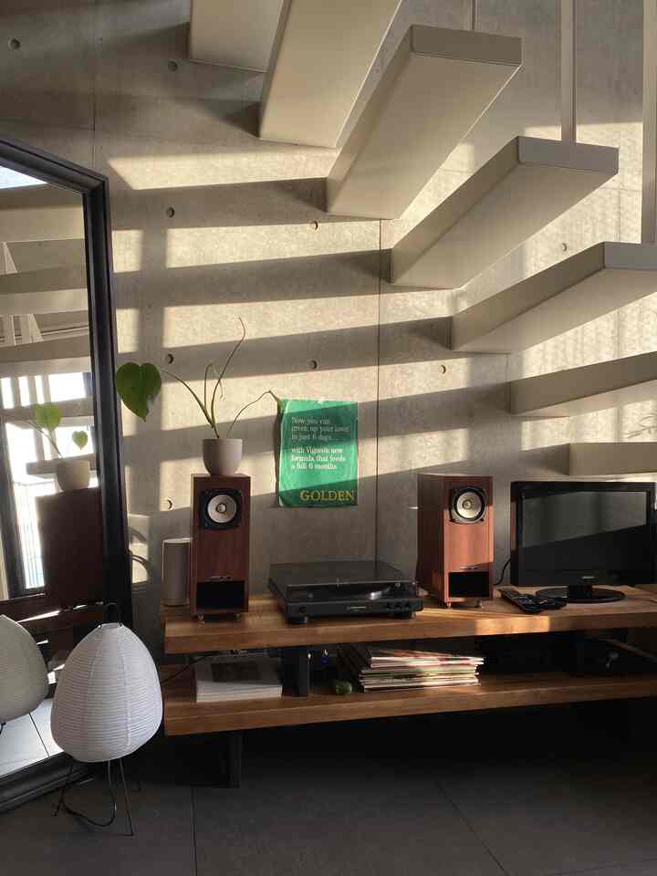 黒い全身鏡と木目調のテレビ台上にスピーカーが置かれたミニマルなコンクリート壁面空間、階段の影が印象的なインテリア