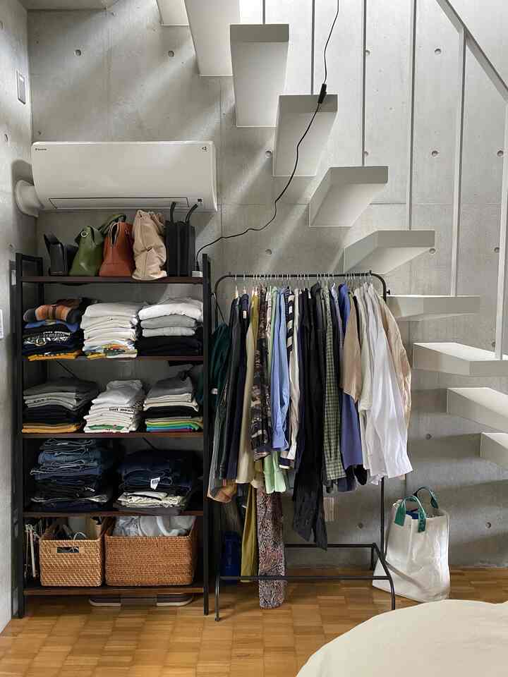 グレーのコンクリート壁と白い階段がある空間に、オープンタイプの服掛けと整然と畳まれた衣服が配置されたすっきりとした収納アイデアのあるインテリア