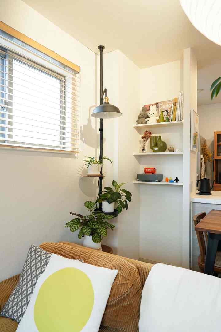 自然光の差し込むリビングのコーナー、ベージュのソファと黒のフロアランプ、効率的に活用されたコーナー棚がある温かみのあるインテリア
