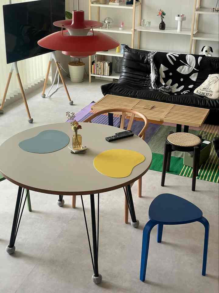 白い壁と木目調の家具があるリビング、黒いソファとスツール、丸いダイニングテーブルに黄色と青のランチョンマットが配置された雰囲気のある空間