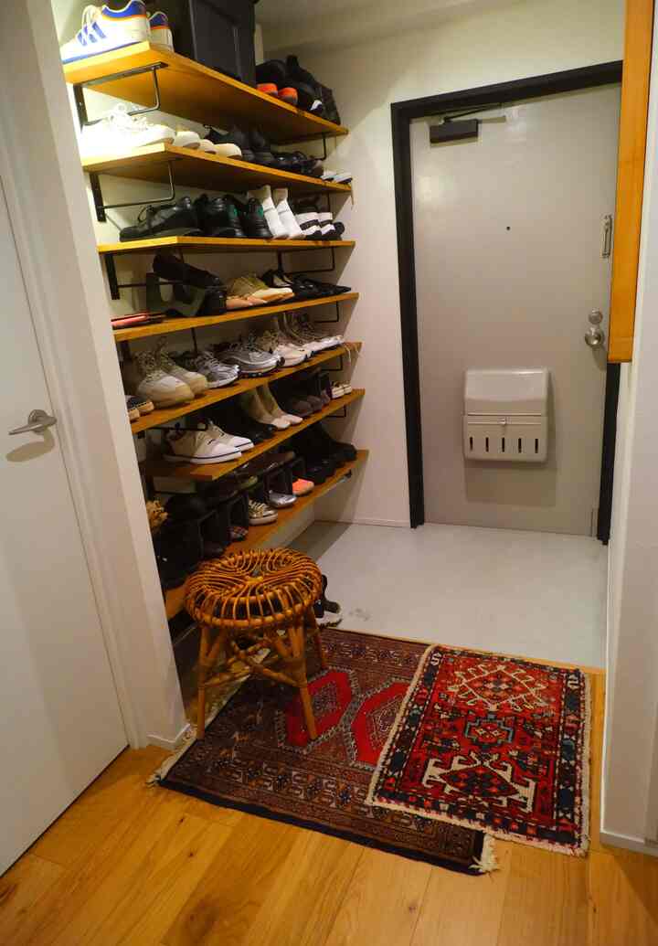 ブラウンのウッドトーンが基調のコンパクトな玄関、靴が並べられた棚、ラタンのスツール、重ねられたヴィンテージラグが配置された雰囲気のある空間