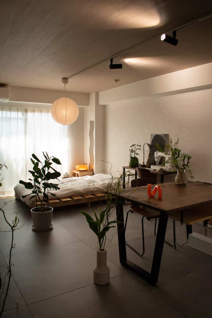 ナチュラルな色調のワンルーム、ベッドと作業スペースが配置された植物と照明のある雰囲気のあるインテリア