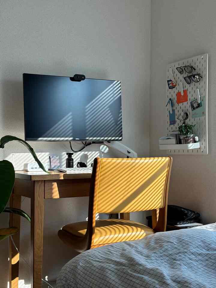白い壁と木目調の家具が調和した小さなホームオフィス、黄色いオフィスチェアと大きなモニターが配置された整った作業空間
