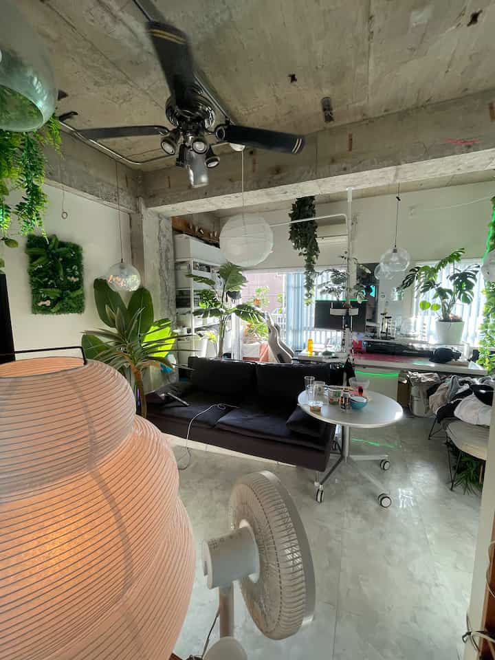 露出したコンクリート天井と黒の3人掛けソファ、豊富な植物とペンダントライトが配置されたナチュラルでインダストリアルなリビング兼ホームオフィス空間