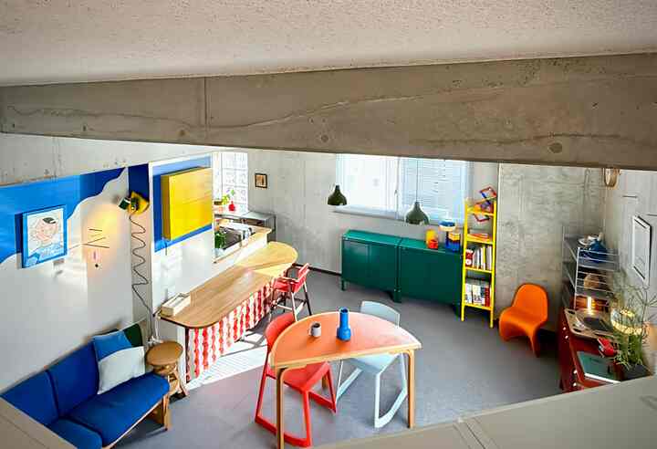 白い壁とグレーの床のモダンなワンルームリビング、青いソファとオレンジのアームチェアが置かれた雰囲気のある空間