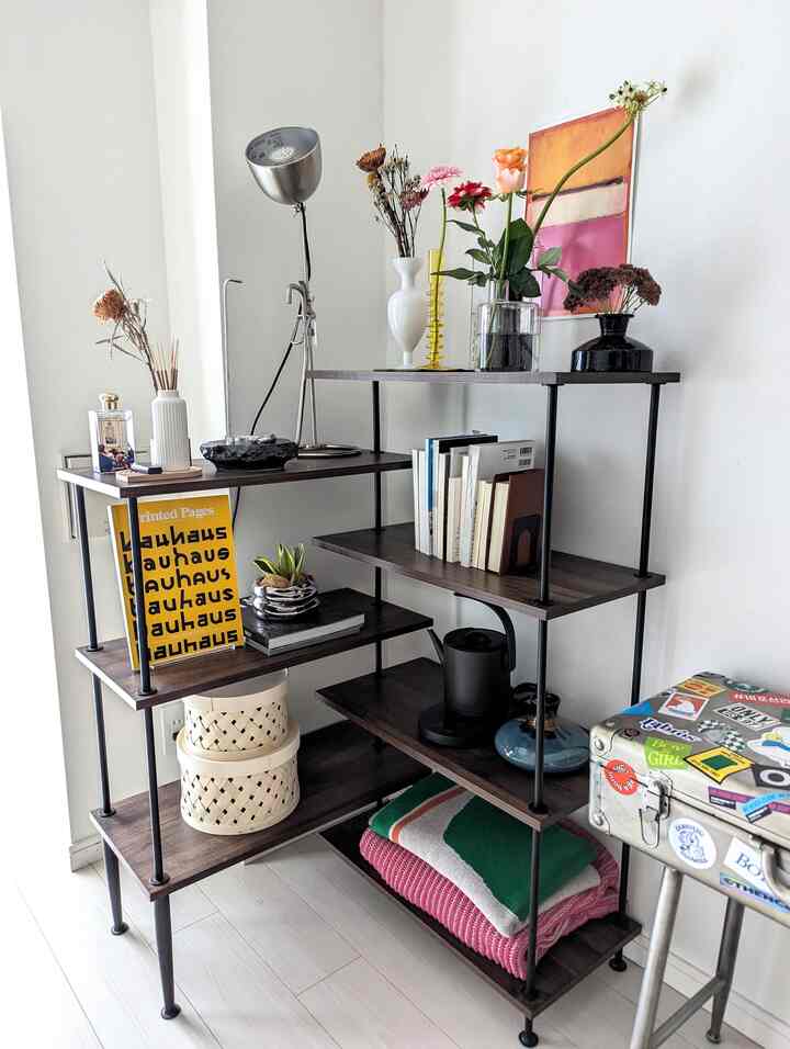 白い壁と床が広がる空間に、ダークブラウンの木製と黒い金属のラックが置かれ、花瓶やポスターなどの装飾品が配されたナチュラルでモダンな雰囲気のインテリア