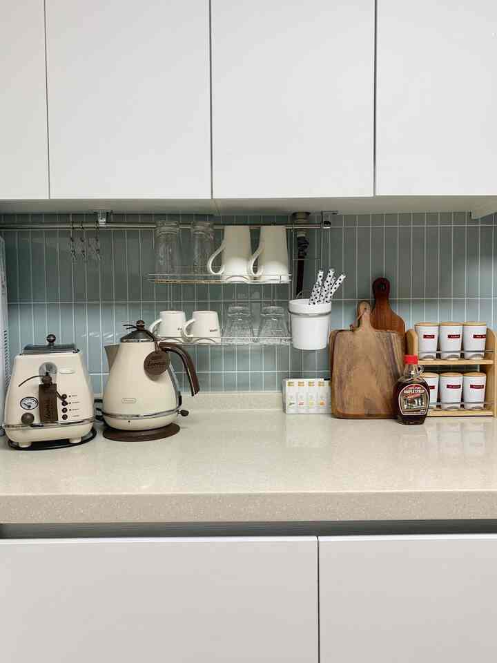 ミント色のタイル壁とベージュの大理石カウンターがあるキッチン、デロンギの電気ケトルとトースター、木製まな板がシンプルに配置された居心地の良いおうちカフェの雰囲気