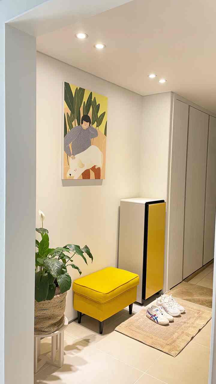 Bright white-walled long entrance featuring a yellow bespoke shoe dresser, matching stool, and a natural plant accent for a cozy atmosphere