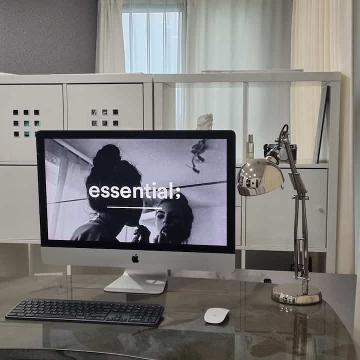 白とシルバーのトーンのワンルームのホームオフィス、チェストとアイマック、作業用ランプが配置された整った空間