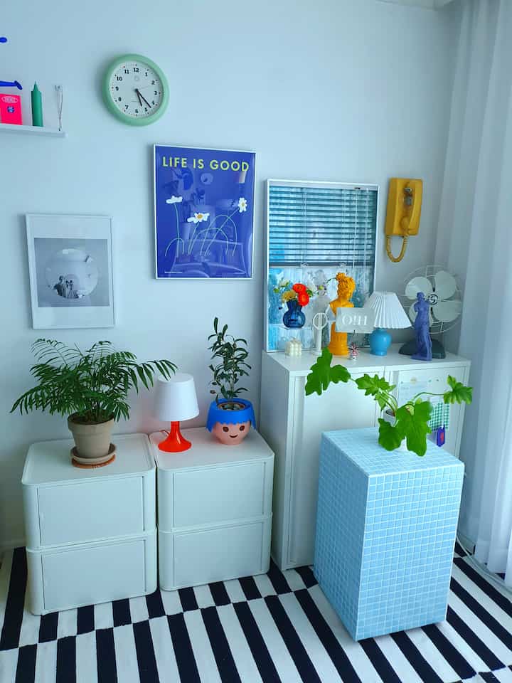 A simple modern studio apartment with white walls and furniture, blue framed poster and tiled cube, featuring various plants