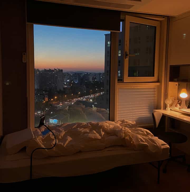 Cozy compact bedroom with white bedding and warm desk lamp, featuring large window overlooking city night view