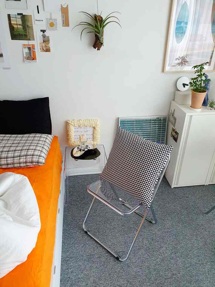 White-walled bedroom with gray carpet floor for a single household, featuring a transparent folding chair with a checkered cushion in a minimal style
