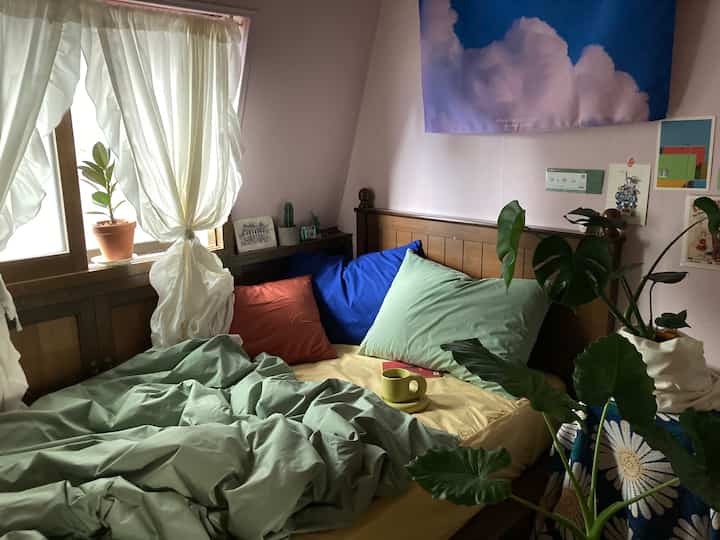 Pastel pink-walled bedroom with wooden bed in green tones, featuring assorted plants and cloud poster creating cozy atmosphere