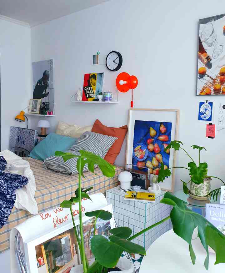 白い壁とベージュの寝具が調和した寝室、大きなモンステラの植物や時計、かわいらしい額縁が配置された雰囲気のある空間