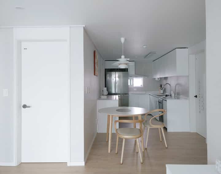 白とナチュラルトーンの縦長キッチン、円形ダイニングテーブルと木製椅子が配置された明るく整った空間