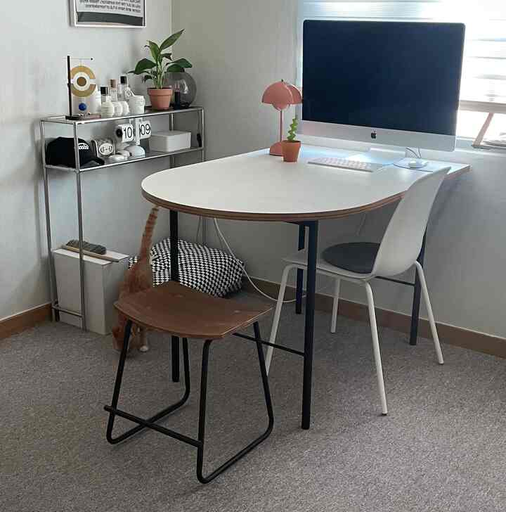 A compact home office space in white and wood tones featuring a desk, chair, stool, shelf, and a cat on the floor