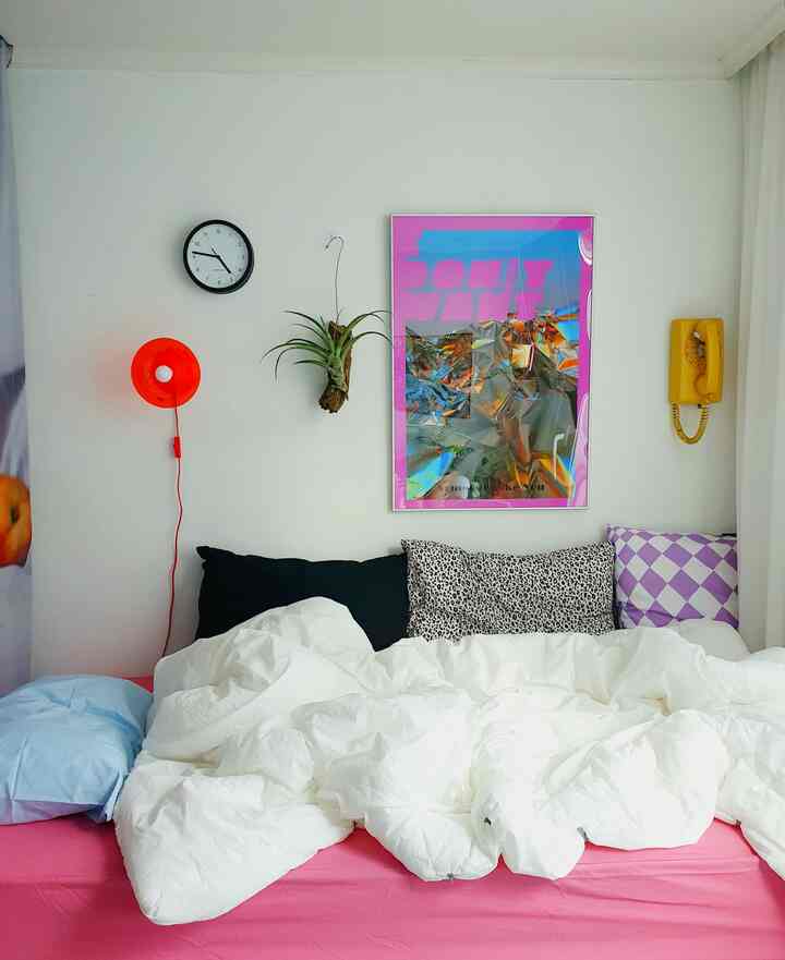 Long and narrow bedroom featuring a pink mattress with white bedding, a colorful poster on the wall, orange wall lamp, and yellow telephone