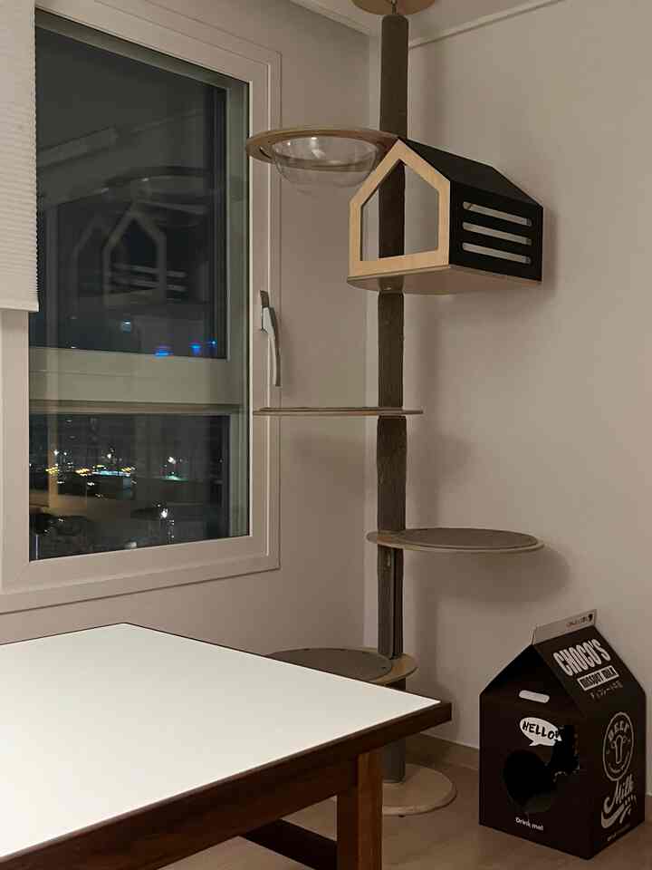 A living room with white and wood tones featuring an Obbyfo wooden cat tower beside the window and a milk carton-shaped cat hideout