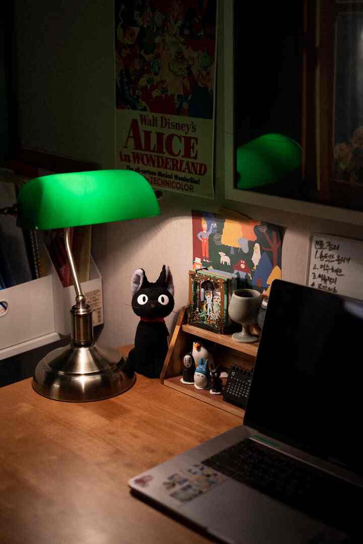 Home office space with warm wood tone desk featuring a green table lamp and Ghibli character objets