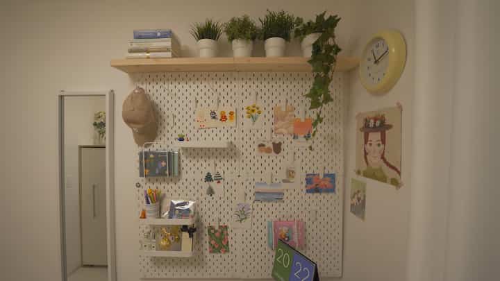 白と木目調が調和した小さな部屋、ウォールシェルフと多彩な壁飾り、黄色い時計が配置された雰囲気のある作業空間