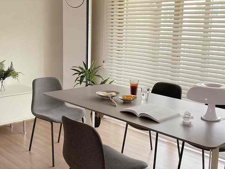 Natural wood-toned living room with a ceramic dining table, blinds, and dining chairs arranged in a cozy setting