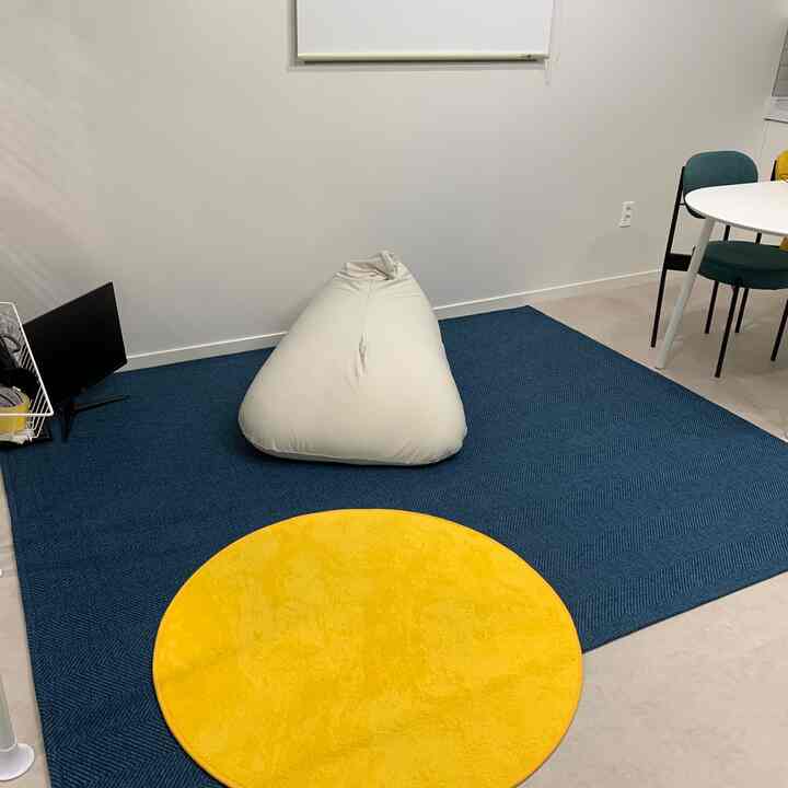 Compact living room with a large blue rectangular rug and yellow round rug, featuring a beige bean bag centrally and a monitor with wire rack on the left side