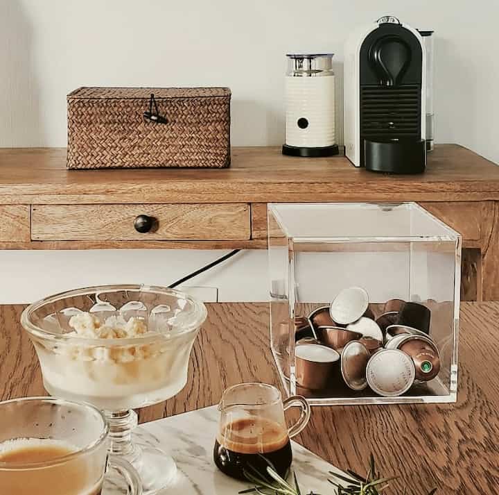 Natural brown toned kitchen space featuring wooden dining table, rattan storage basket, Nespresso coffee machine and capsule container in a cozy home cafe setting