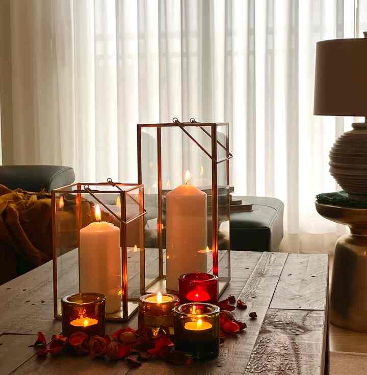 A warm living room with soft beige curtains in the background, featuring gold frame candle holders and cozy lit candles on a wooden side table