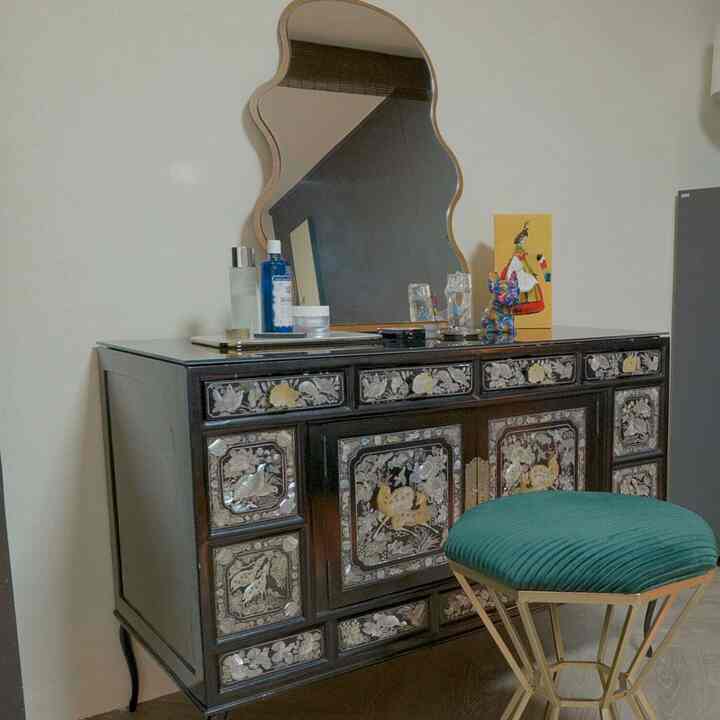 ゴールドフレームの波型鏡とグリーンベルベットスツールが配置された、伝統模様の黒い化粧台がある落ち着いたインテリア空間