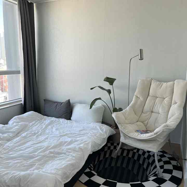 Natural-toned bedroom featuring white bed, armchair, reading lamp, and large indoor plant creating cozy atmosphere
