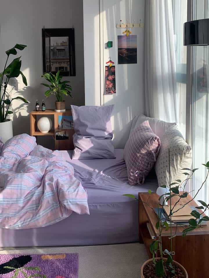 A cozy bedroom with purple bedding, white curtains, plants, and wooden nightstand illuminated by natural light