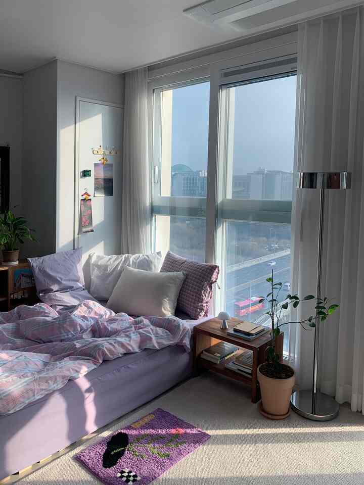 A white and lavender toned bedroom with large window, side table, floor lamp, and plants creating a cozy atmosphere