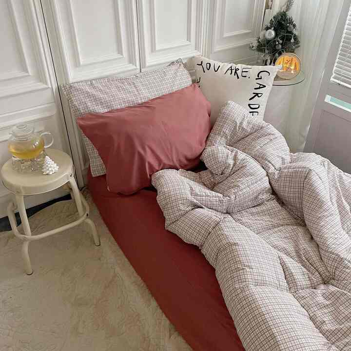 ベージュと赤の色調が調和した寝室、チェック柄の掛け布団とクッションが置かれ、ミッドセンチュリーモダンスタイルのスツールが配された印象的な空間