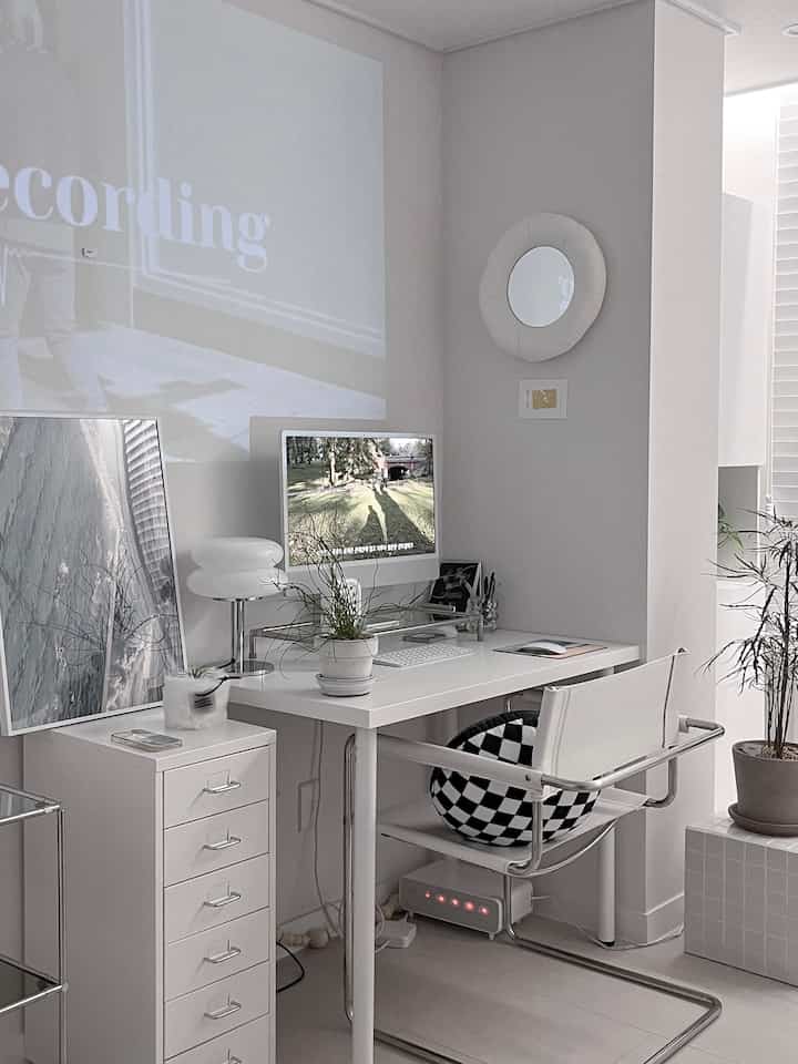 White-toned home office featuring a white desk, chair, monitor stand, and plants in a clean minimal atmosphere