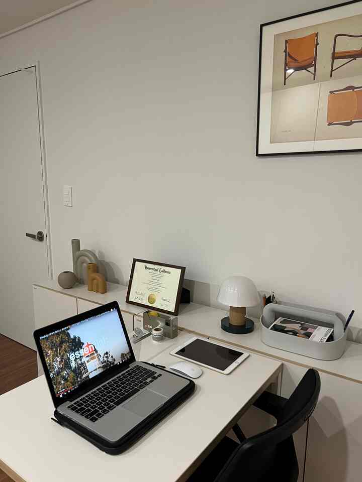 A white-walled study room with brown flooring featuring a clean white desk, laptop, and a modern table lamp in a contemporary home office setup
