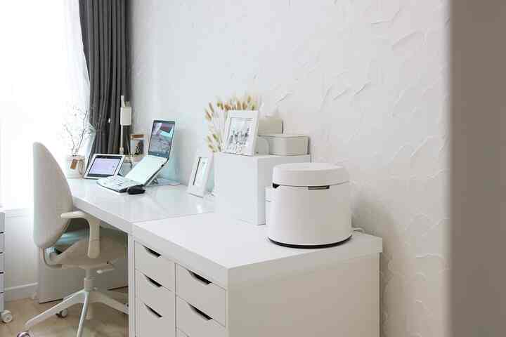 White-toned clean home office featuring a long desk and drawer unit with laptop, picture frames, and vase in a modern workspace
