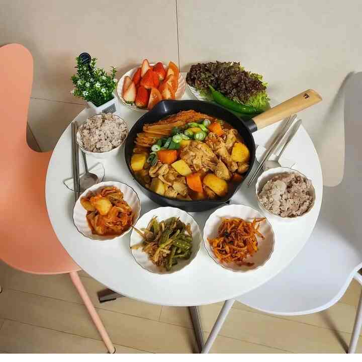 白とピンクのダイニングチェアが配置された丸い白いテーブルのある清潔感のある二人用ダイニングスペース、素朴な鶏煮込みとおかずが並ぶ暖かい食事シーン