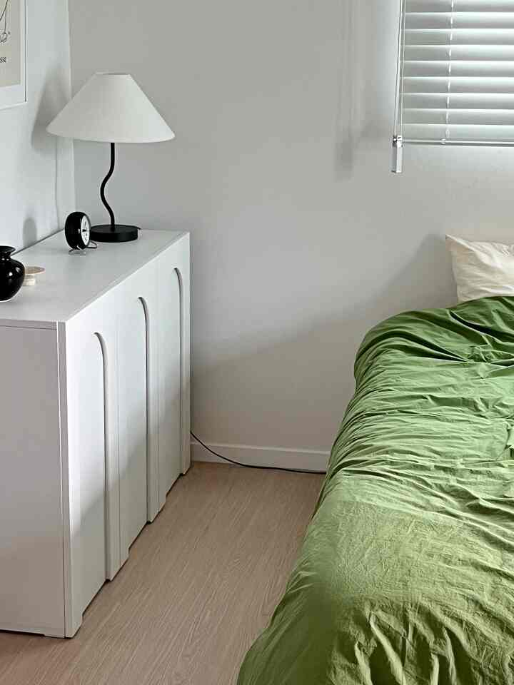 A bedroom with white and natural wood tones, featuring a green bedspread and stylish storage cabinet providing a cozy atmosphere
