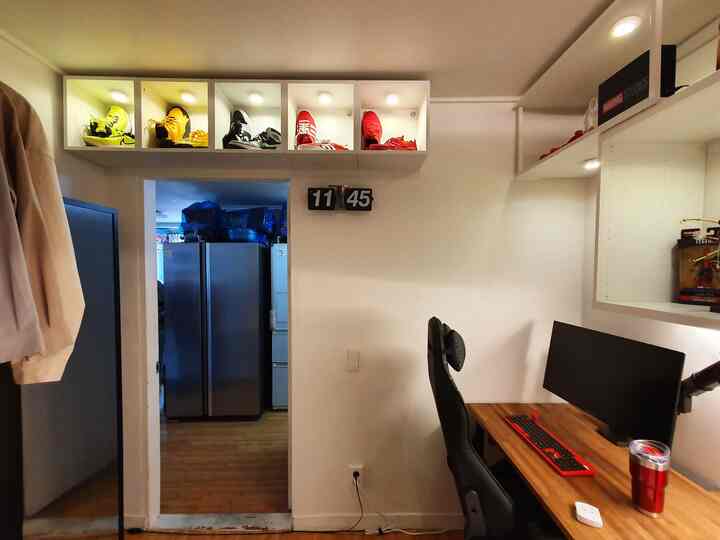 A compact, practical home office space with white walls and wood-tone desk, featuring wall shelves and a flip clock