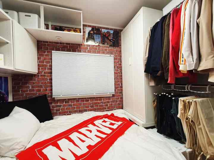 Small bedroom with brick-patterned walls and white wardrobe, featuring a white bed with a red Marvel blanket centrally placed