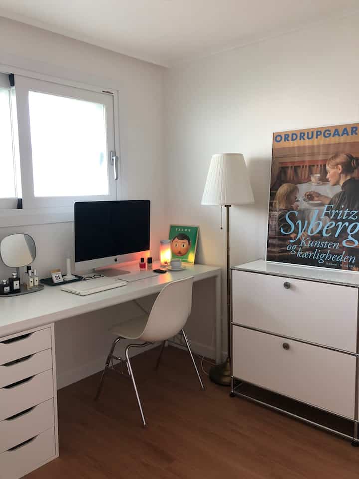 白い壁と木目調の床が特徴的な書斎、デスクとチェア、フロアランプ、壁に掛かった額縁が配置されたモダンで清潔感のあるインテリア