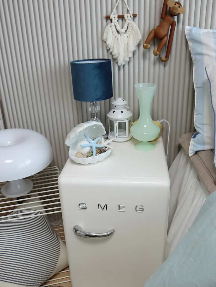 White and blue toned bedroom corner featuring a mini fridge nightstand with small lamps and decorative items, creating a cozy atmosphere