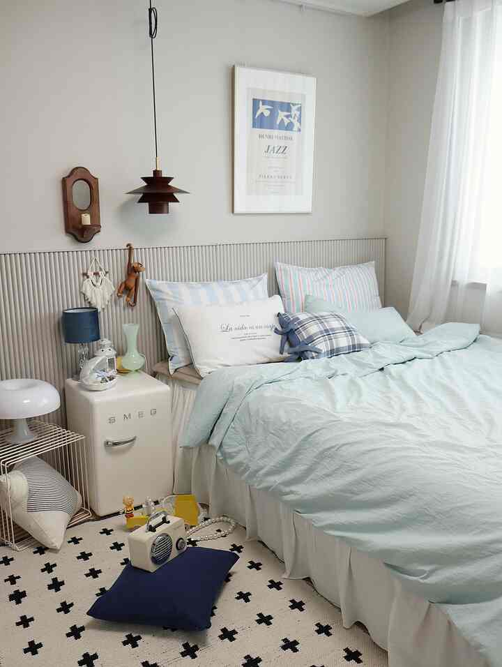 パステルトーンの青い寝室、ストライプ柄の枕やクッション、ナイトスタンドと照明が配置された雰囲気のある空間