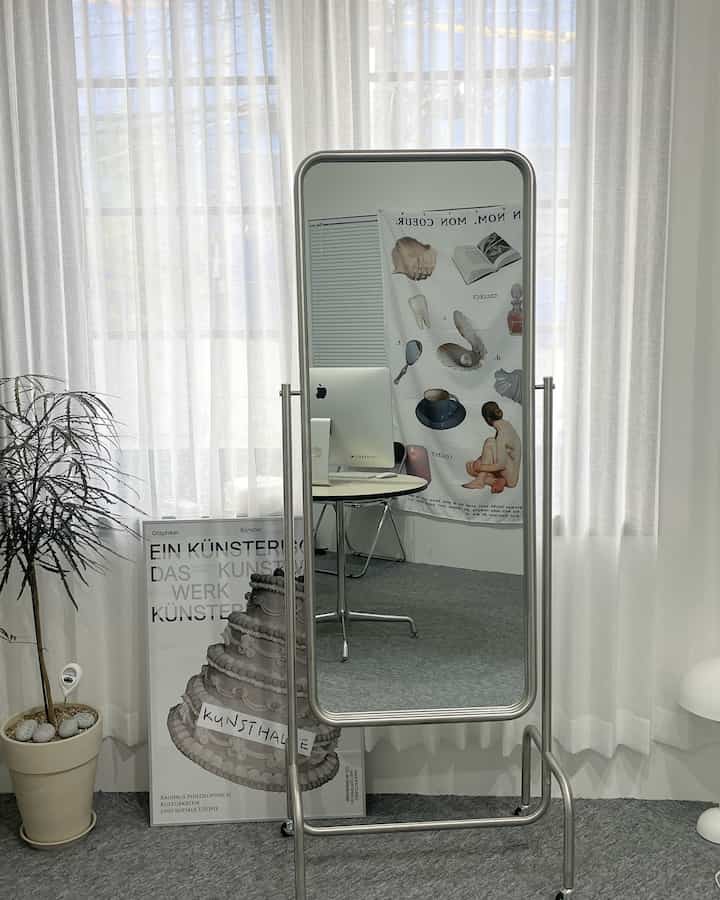 ホワイトトーンのミニマルなリビング、スタンドミラーに映る丸テーブルとパソコンが配置されたシンプルな空間