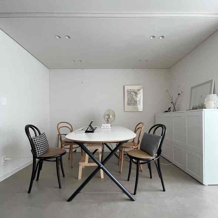 A white-toned dining room and home office space featuring a white table, black and oak chairs, framed artwork, and cushions in a clean modern interior