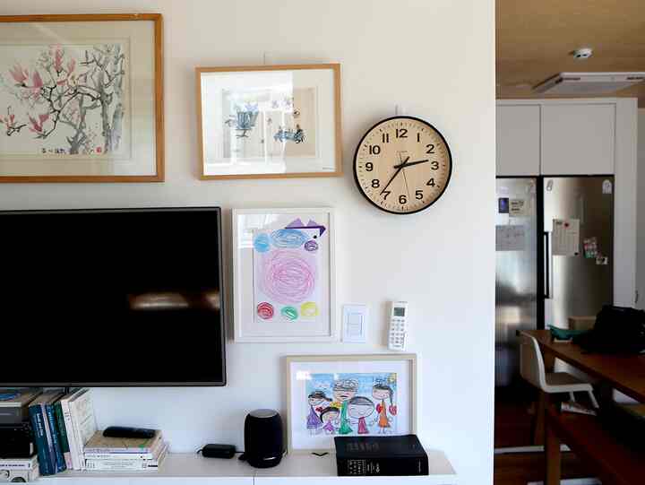 A white wall living room featuring wooden framed picture frames, a large numbered wall clock, and a mounted TV in a simple setting