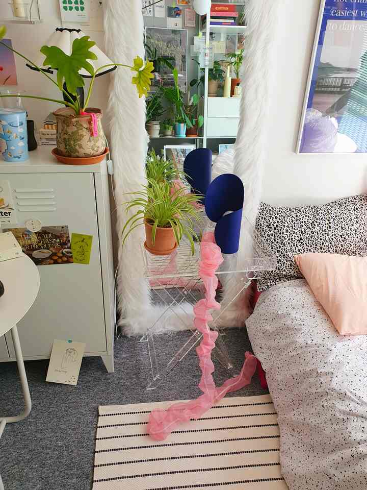 A modern bedroom in white and gray tones featuring a full-length mirror, transparent table, and various plants creating a cozy atmosphere