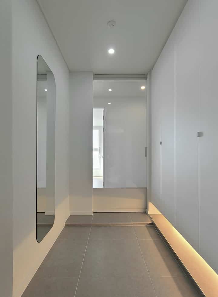 Modern entrance with white and gray tones featuring a shoe cabinet along the wall, a tall full-length mirror, and a sliding glass door in a clean setting