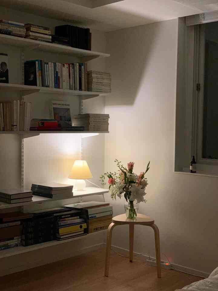 A simple, modern study and living room with warm lighting, featuring wood tone stool and white wall shelves creating a cozy atmosphere