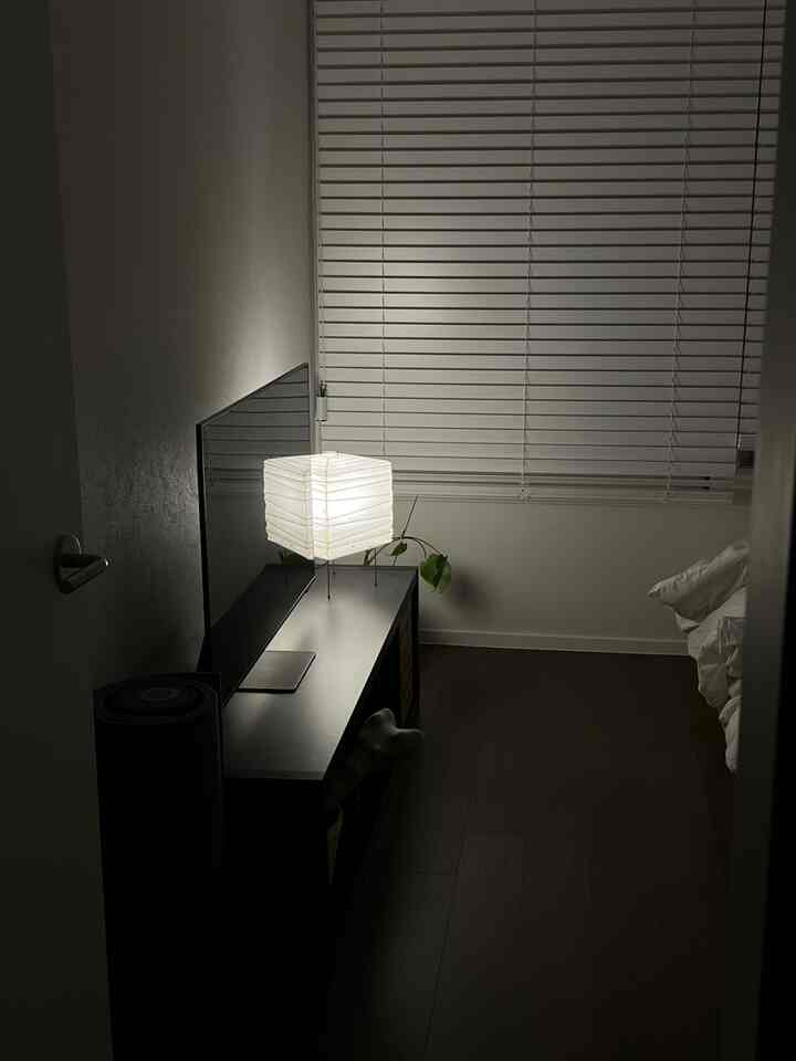 Bedroom with dark wood tone flooring and white blinds, featuring a bright Akari lamp at center-left creating a cozy atmosphere